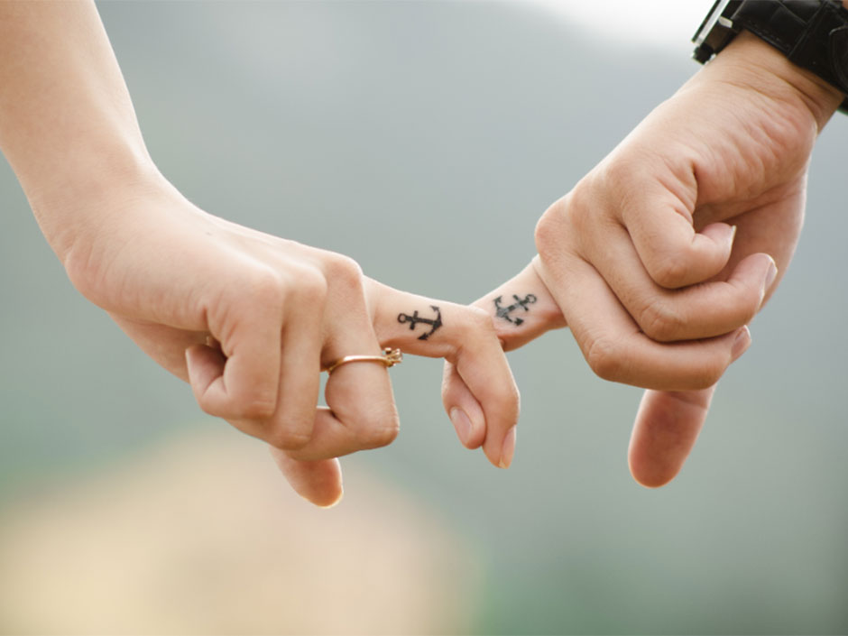 Zwie Hände halten sich an den Zeigefingern, die Finger haben jeweils ein Ankertattoo
